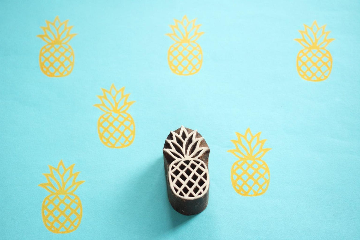 Hand carved Indian wooden printing block with a pineapple design shown on a blue surface with repeated yellow pineapple prints.