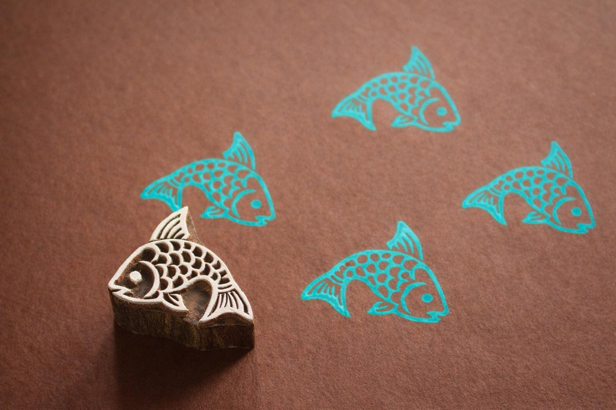 Hand carved Indian wooden printing block with a fish design shown on brown paper with repeated turquoise fish prints.