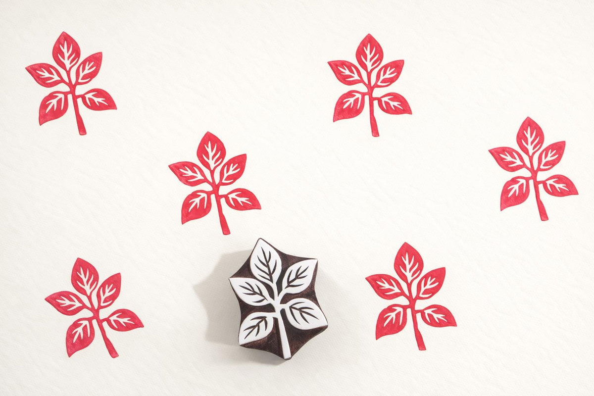 Hand carved four-leaf wooden block printing stamp creating repeating red botanical motif pattern on cream fabric.