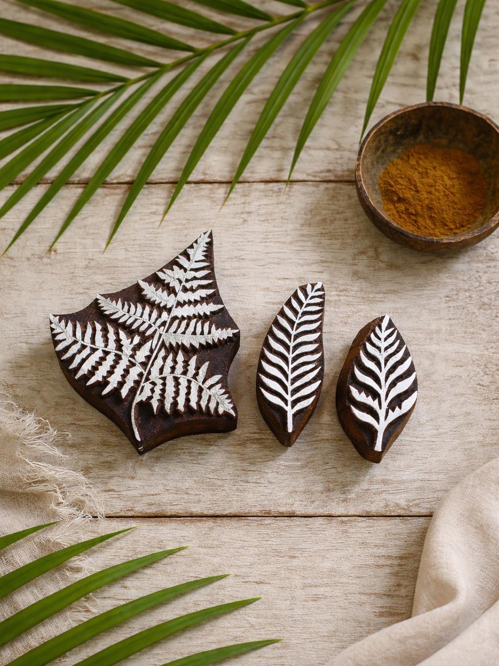 Set of three hand carved wooden fern leaf block printing stamps displayed on rustic wooden surface with palm leaves and bowl of turmeric.