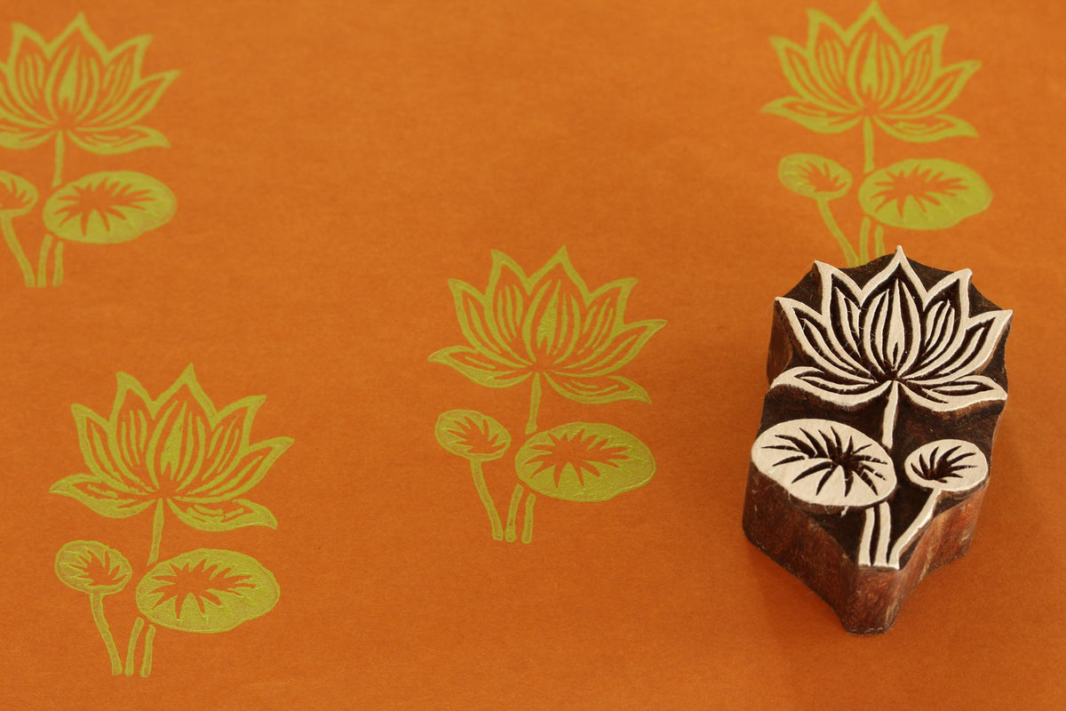 Hand carved Indian wooden printing block with a detailed lotus flower design shown on orange paper with repeated lotus prints.