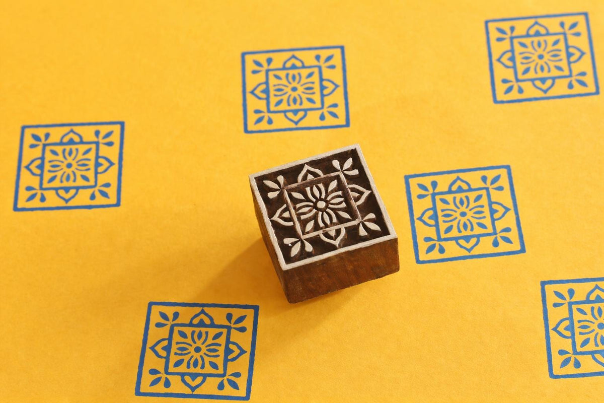 Hand carved square floral wooden printing block on yellow background with blue printed tile pattern impressions.