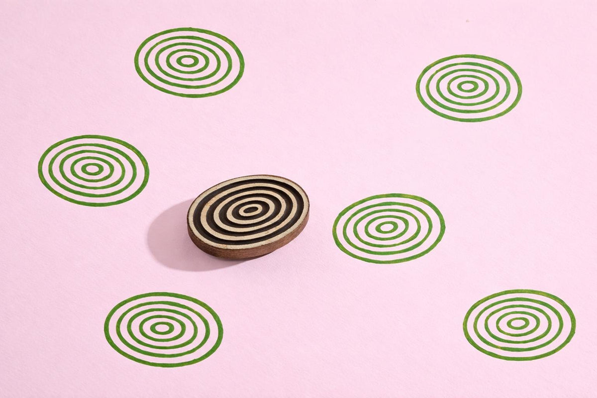 Oval wooden block printing stamp with concentric circle design creating repeating green ripple pattern on pink background.