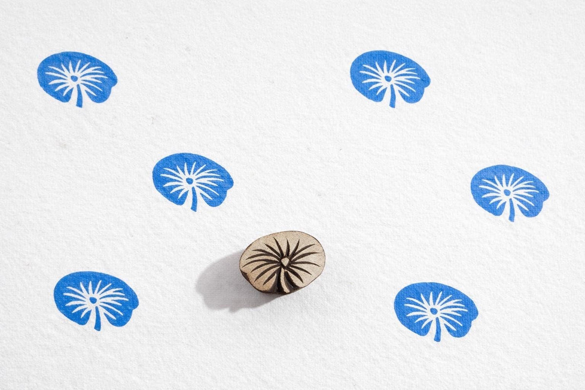Small teardrop shaped lily pad wooden printing stamp making repeating blue leaf pattern on white background.