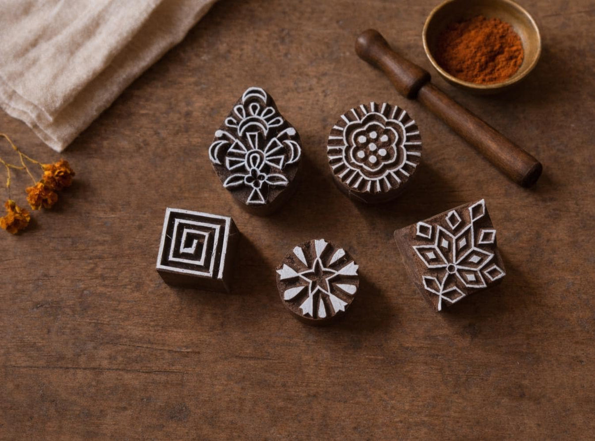 Five hand carved wooden block printing stamps on a wooden table showing Indian textile motifs including a spiral square, sunburst flower, floral rosette, geometric leaf motif, and ornamental folk design.