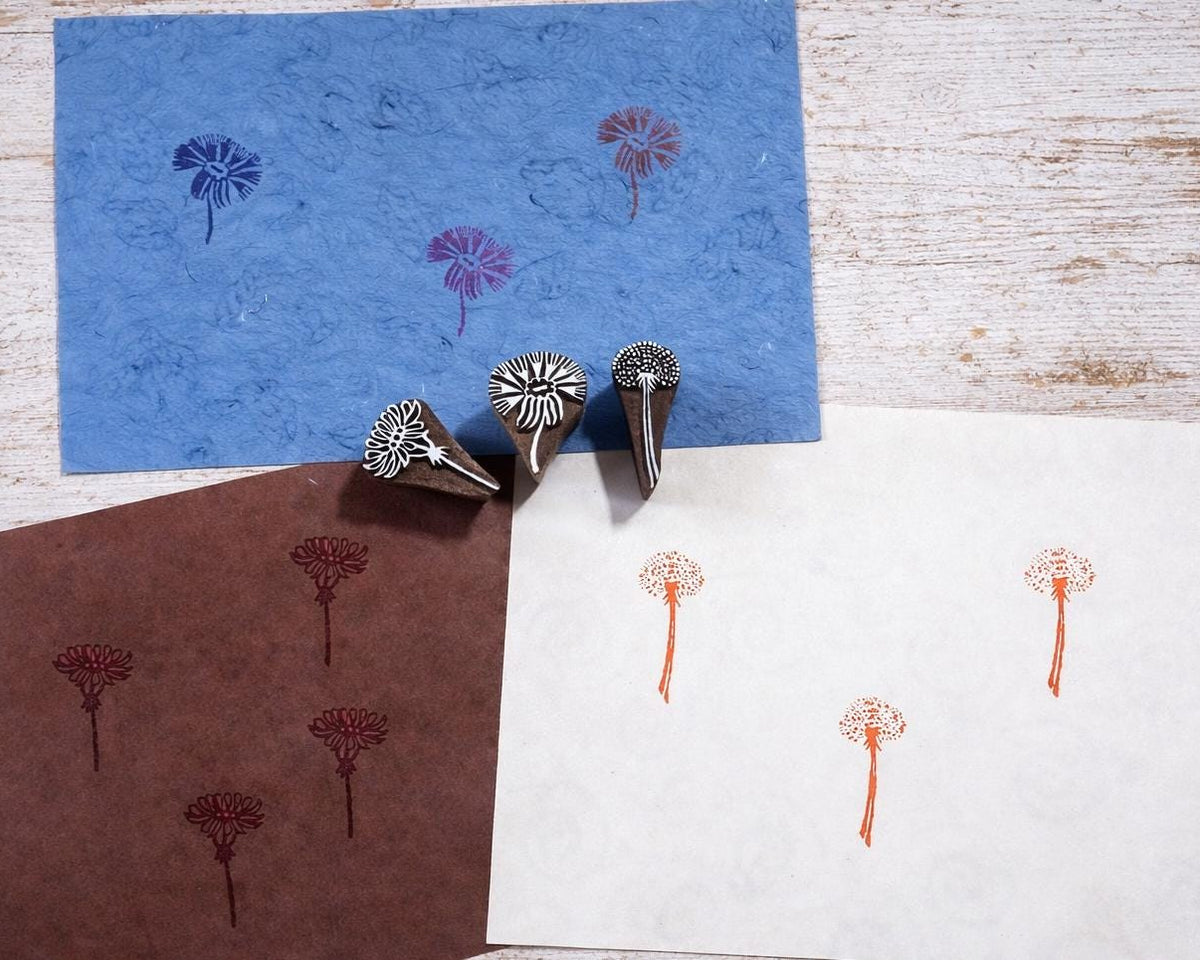 Three hand carved wooden wildflower block printing stamps displayed with printed botanical patterns on blue, brown and white paper.