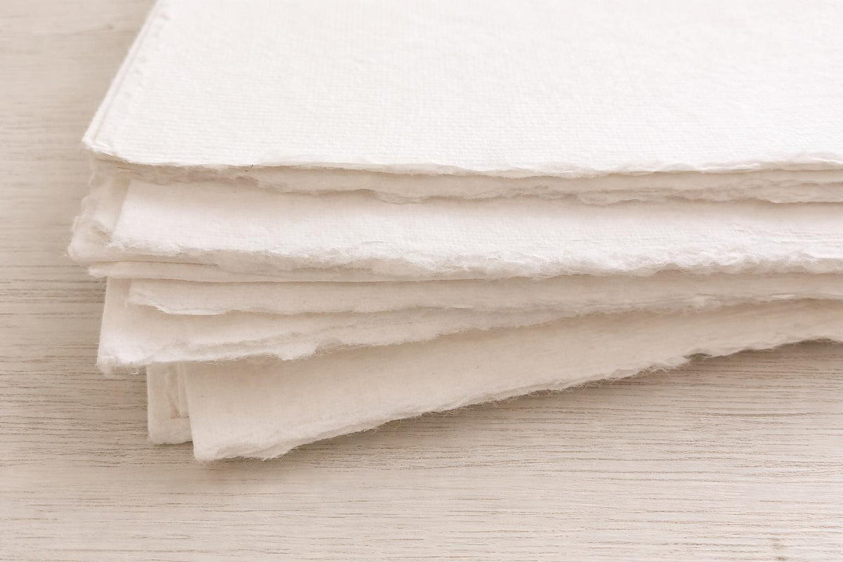 Stack of handmade off-white deckle edge paper sheets viewed from the side, highlighting rough torn edges and thick textured cream paper