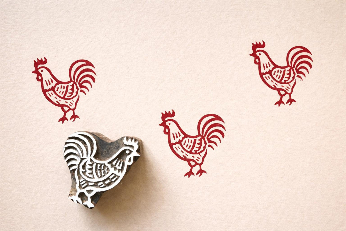 Hand-carved wooden rooster block stamp with red chicken prints on paper in a rustic folk art style