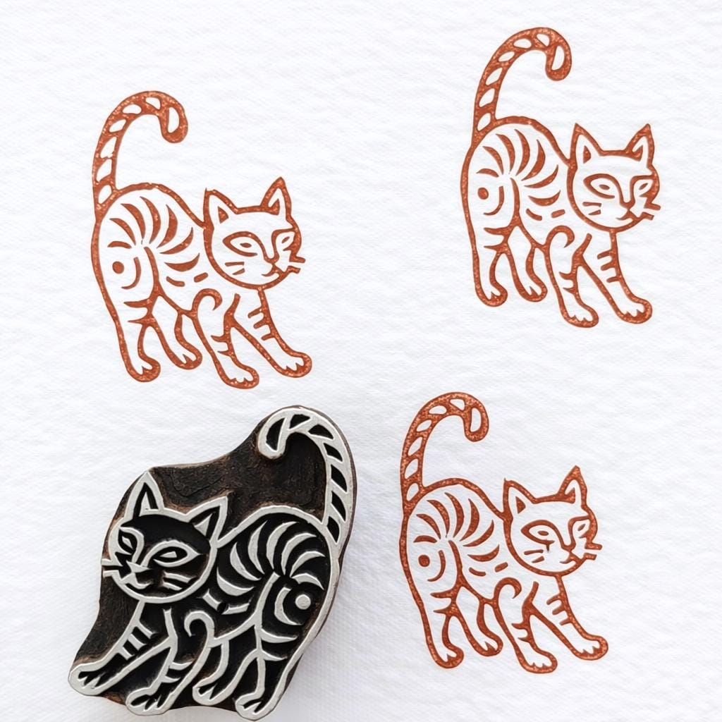 Hand carved walking cat wooden block printing stamp creating burnt orange cat prints on white fabric paper background.