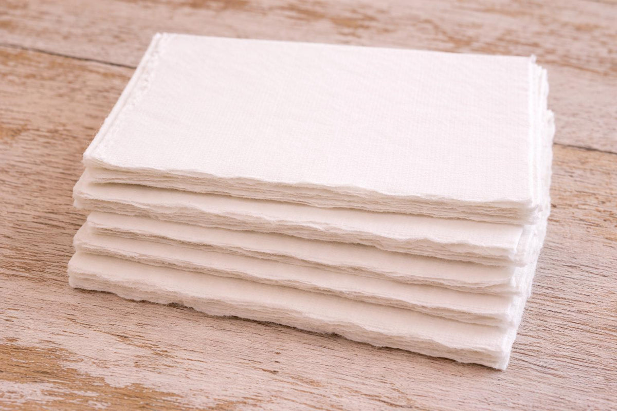 Single off-white handmade paper sheet with deckle edges laid flat on a wooden table, showing soft cream color and textured surface