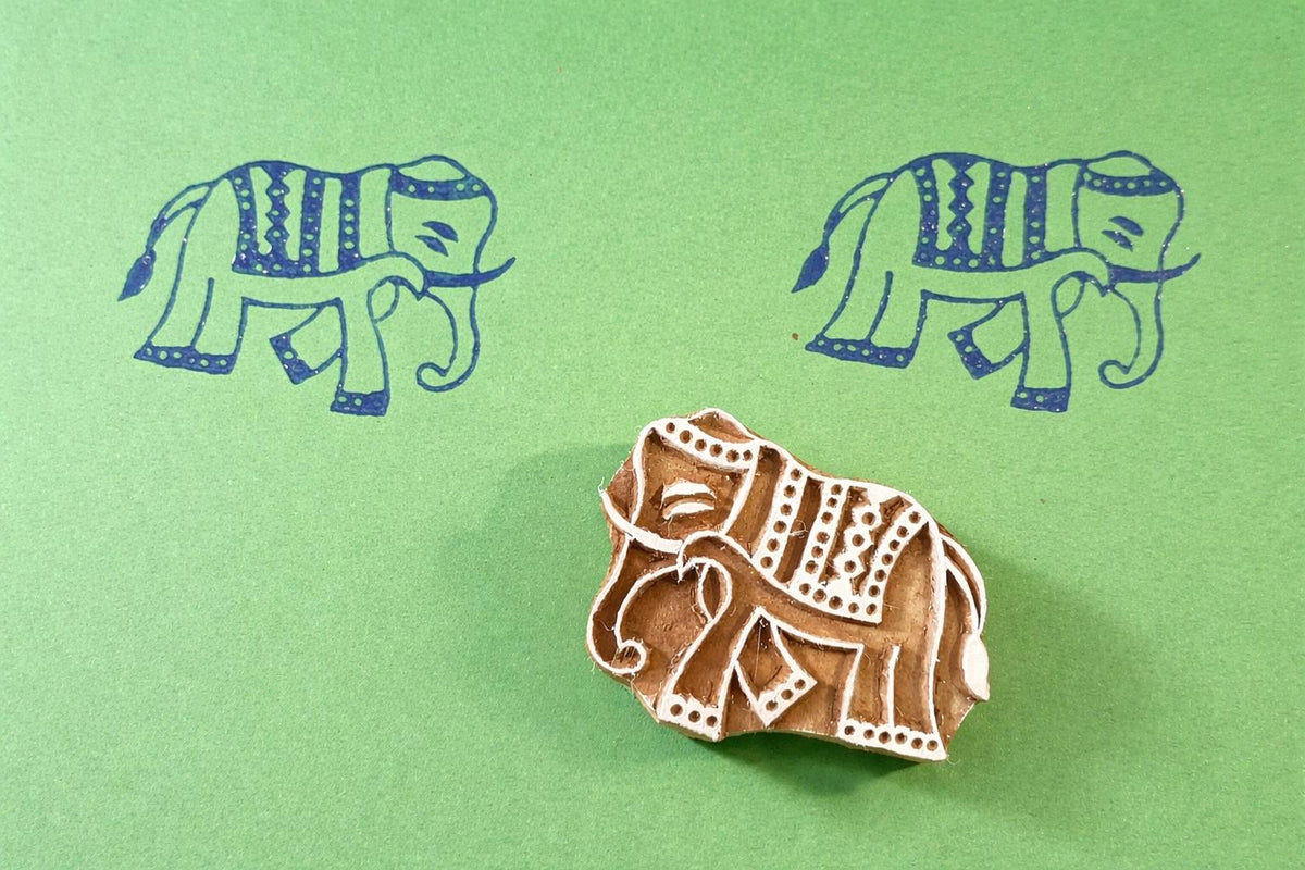 Hand-carved wooden elephant stamp with decorative boho pattern, shown printing repeating elephant designs on green paper.