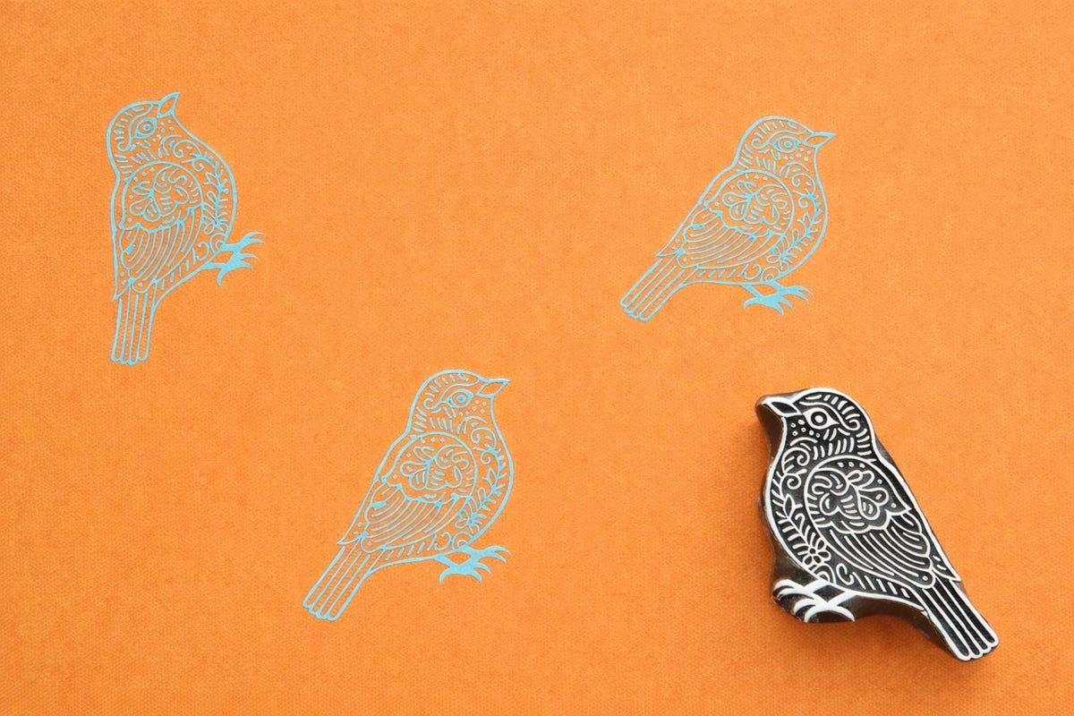 Hand carved wooden block stamp of a small decorative bird with stamped blue bird pattern on orange paper