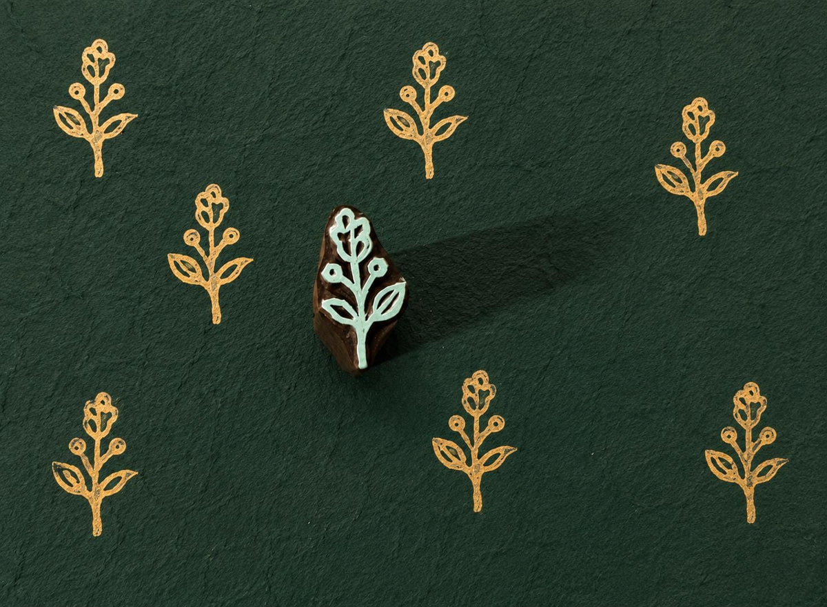 Tiny hand carved wooden block stamp with stylized folk bud sprig motif printed repeatedly on dark textured handmade paper.