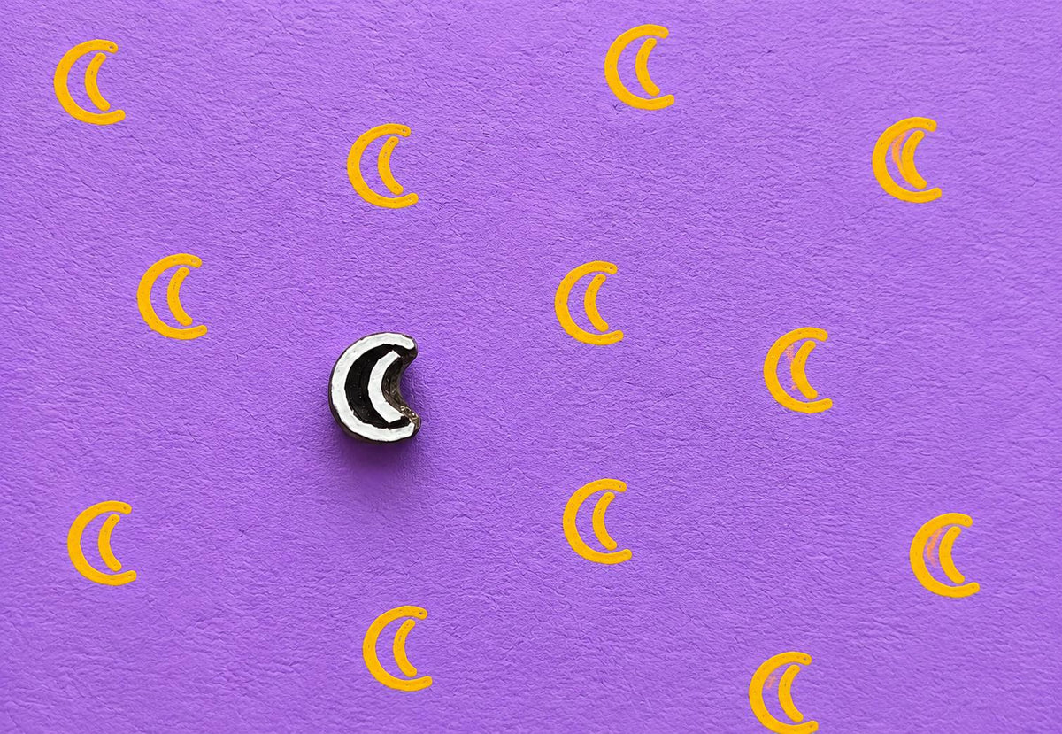 Tiny hand carved wooden crescent moon block stamp printing repeating moon motifs in golden yellow ink on textured lavender paper