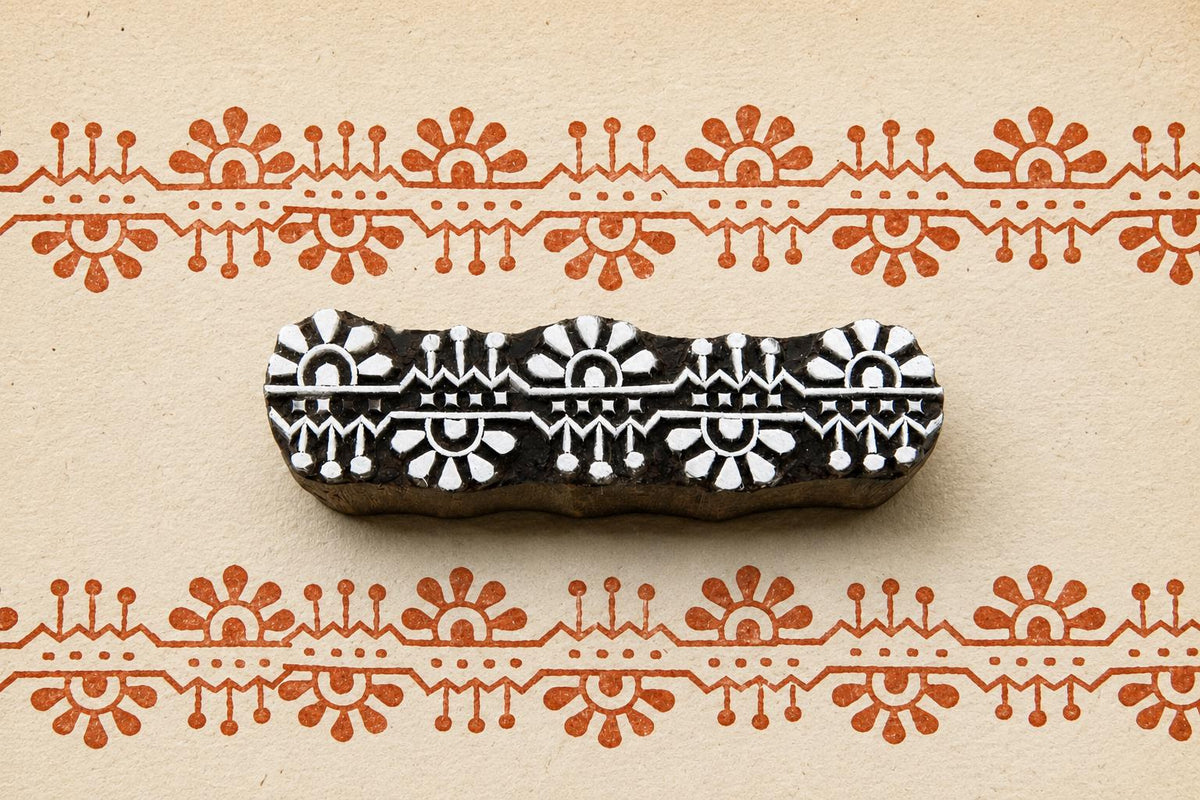 Hand carved wooden border printing block featuring repeating sunburst and geometric folk motifs stamped in terracotta ink on handmade paper.