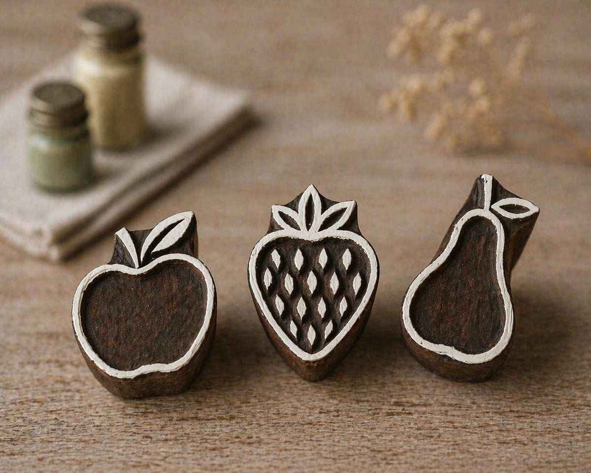 Set of three hand carved wooden fruit printing blocks featuring apple, strawberry, and pear designs arranged on rustic linen fabric background for textile printing and craft projects.