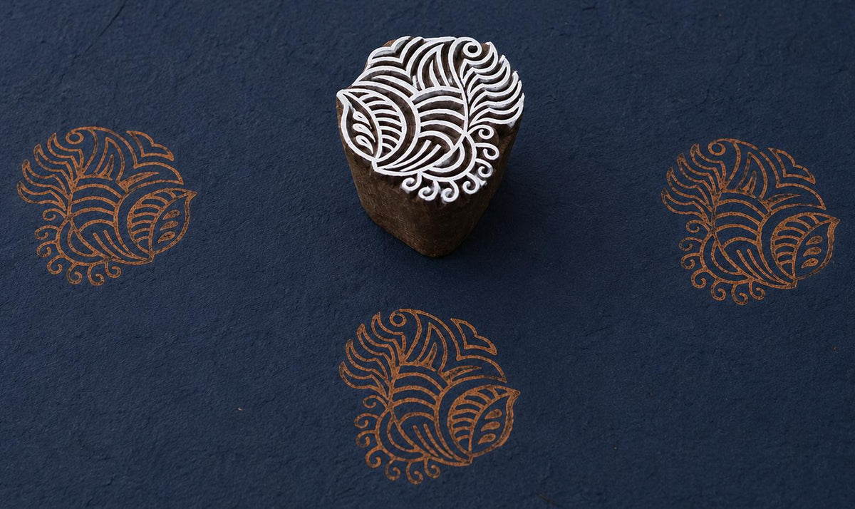 Hand carved Indian wooden printing block featuring an ornate paisley leaf motif stamped in copper ink on deep navy handmade paper, suitable for fabric printing, clay work, and textile design.