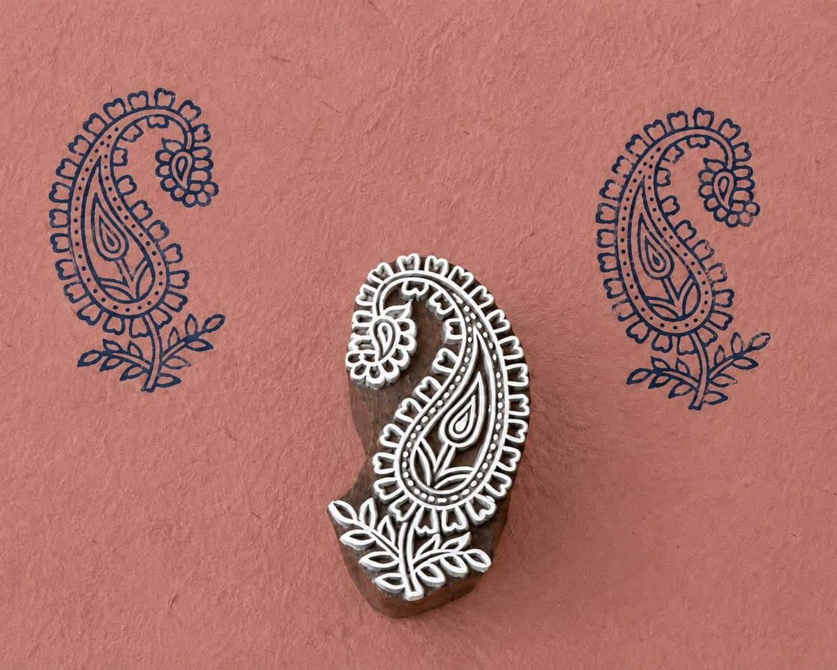 Hand-carved wooden printing block featuring a paisley flower motif with leaves, stamped in navy blue on textured terracotta handmade paper.