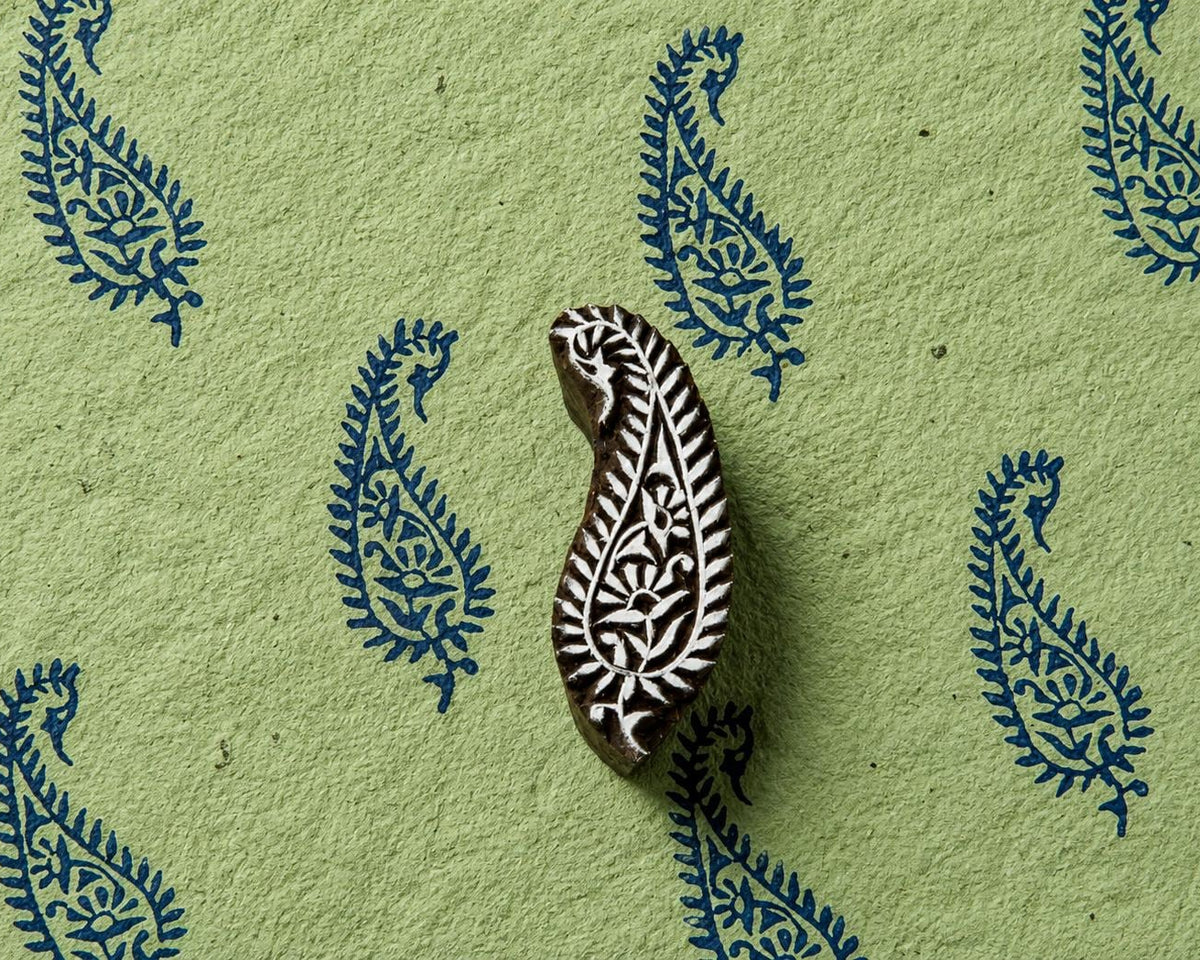 Hand carved wooden paisley printing block displayed with repeating blue floral paisley impressions on textured handmade paper.