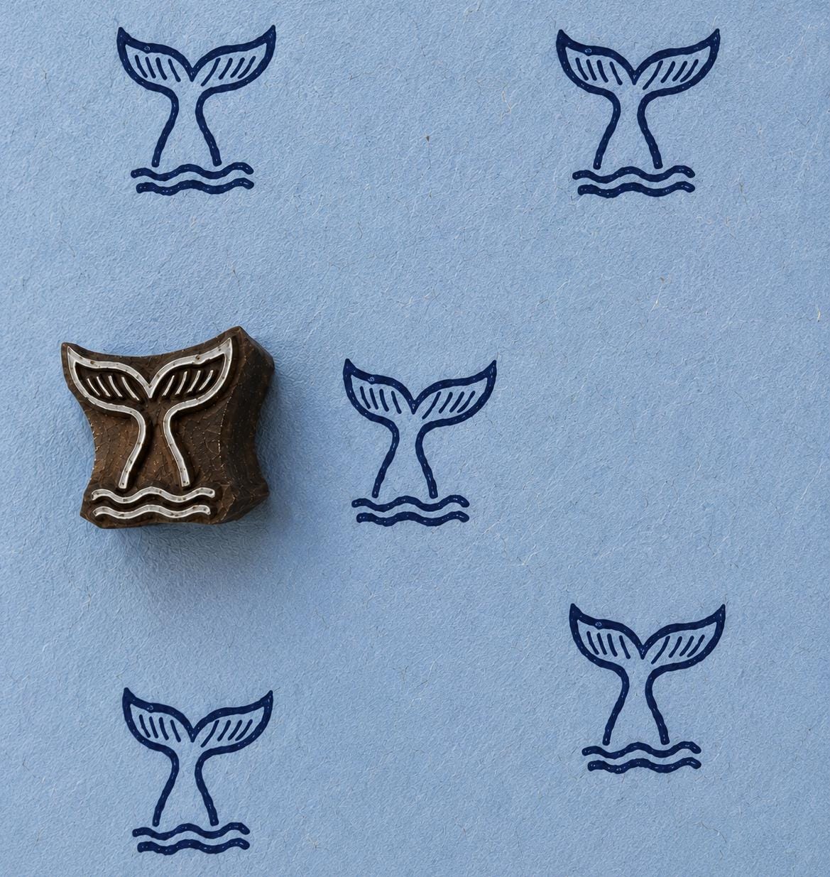 Hand carved whale tail wooden block stamp displayed on blue handmade paper with repeating ocean-inspired impressions.