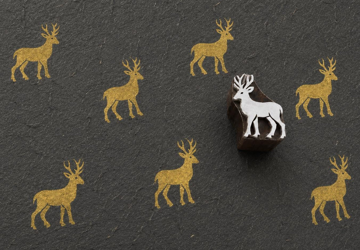 Hand-carved wooden printing block featuring a standing buck deer with branching antlers stamped in metallic gold on textured black handmade paper.