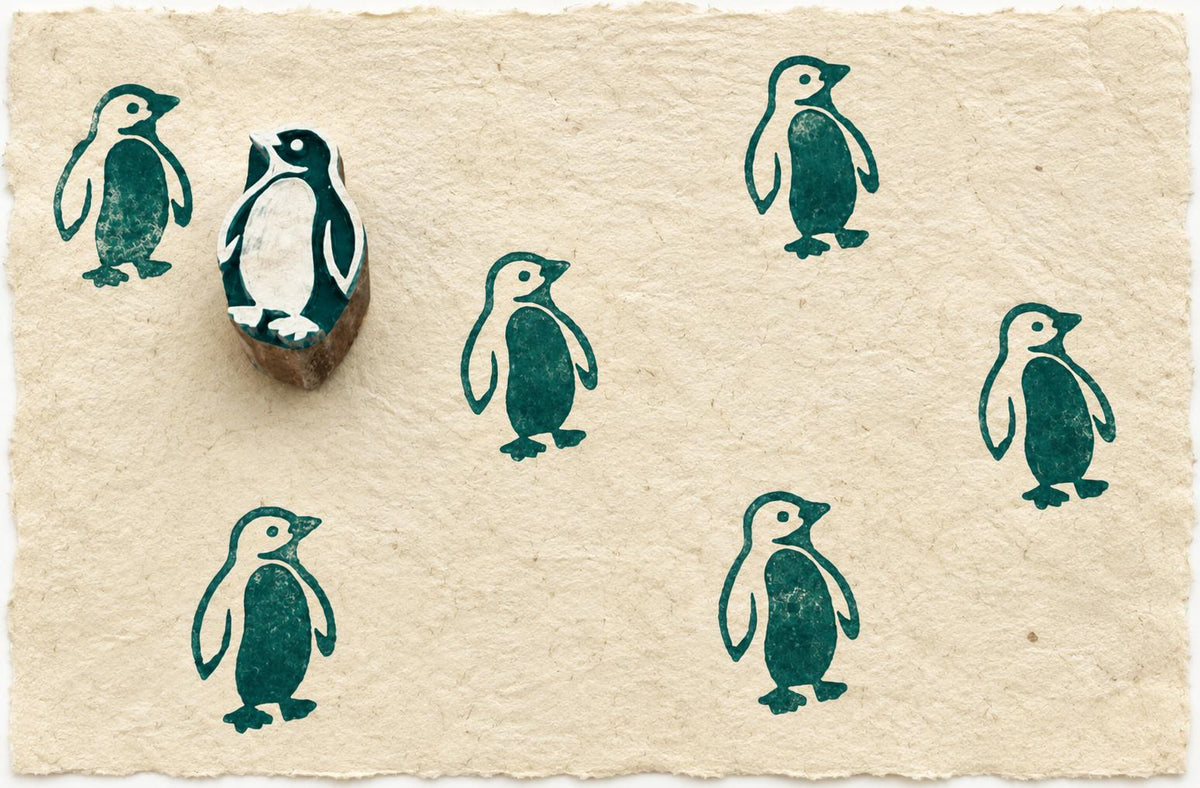 Hand carved penguin wooden block stamp displayed on handmade paper with repeating teal penguin prints.