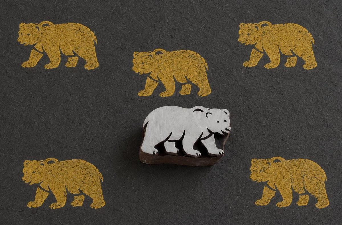 Hand-carved rustic bear wooden printing block displayed on dark handmade paper with repeated golden bear impressions.