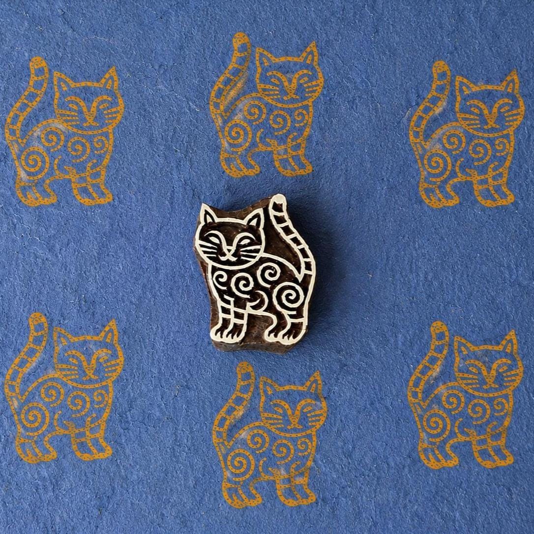 Hand-carved wooden stylized cat printing block with repeated ochre cat impressions on blue handmade paper, traditional Indian textile stamp.
