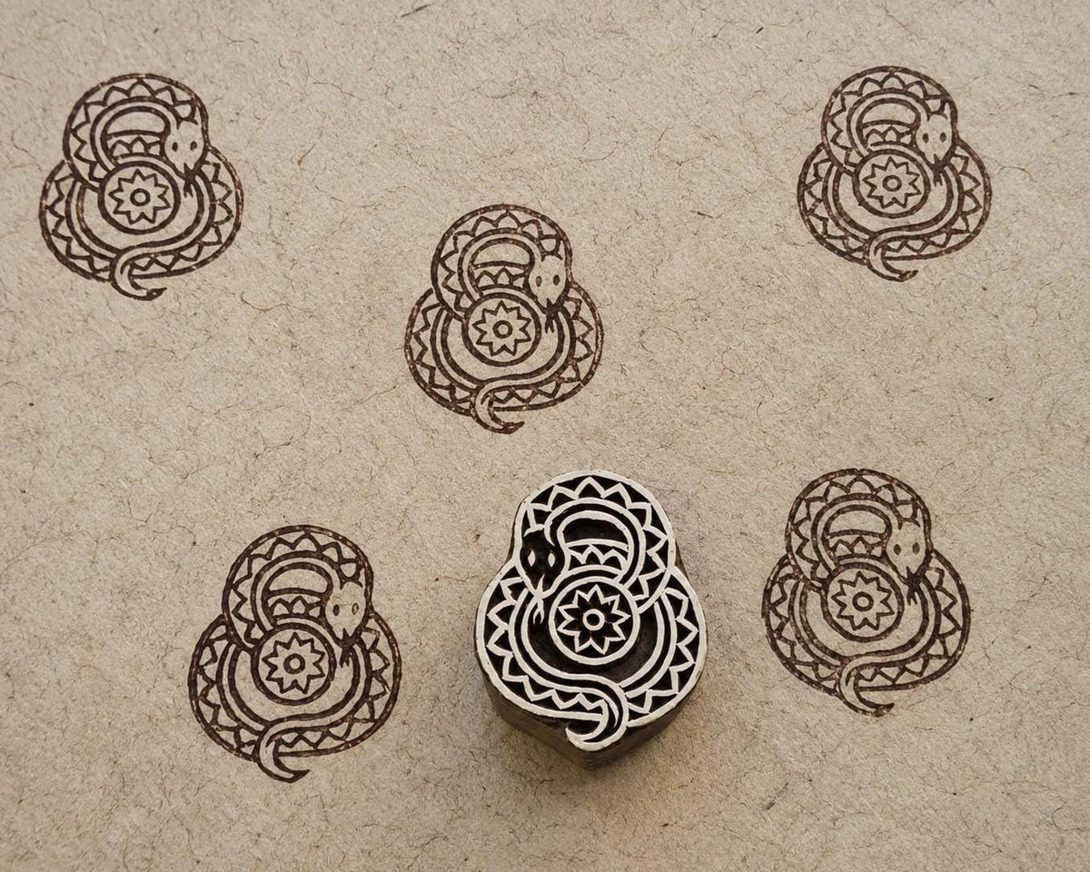 Hand-carved wooden folk art snake printing block with repeated brown coiled snake impressions on handmade paper.