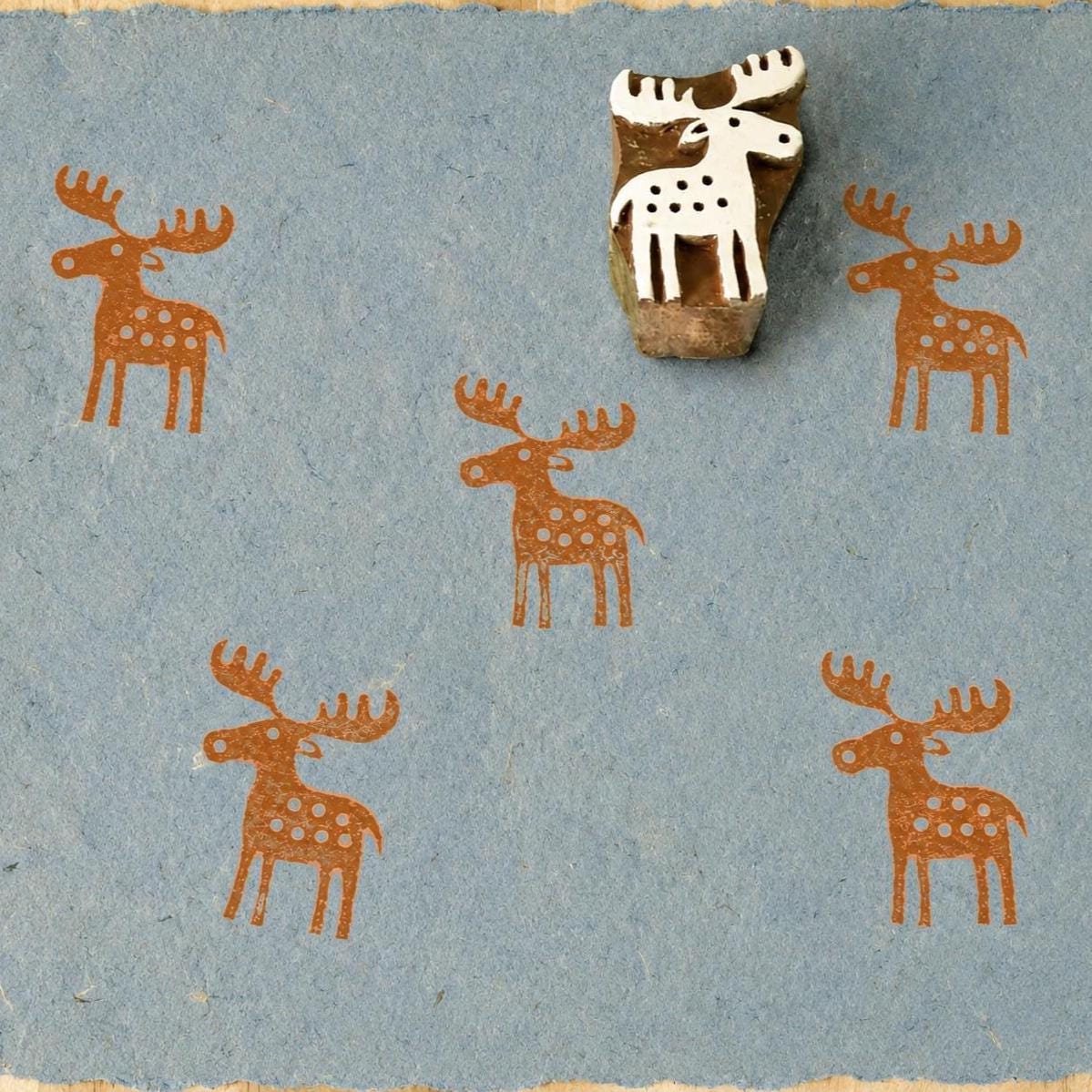 Hand-carved wooden folk art reindeer printing block with repeated terracotta reindeer impressions on blue handmade paper.