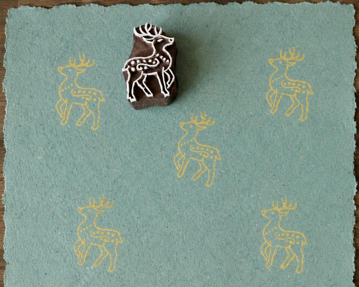Hand-carved wooden woodland deer printing block with repeated pale yellow deer impressions on blue handmade paper.