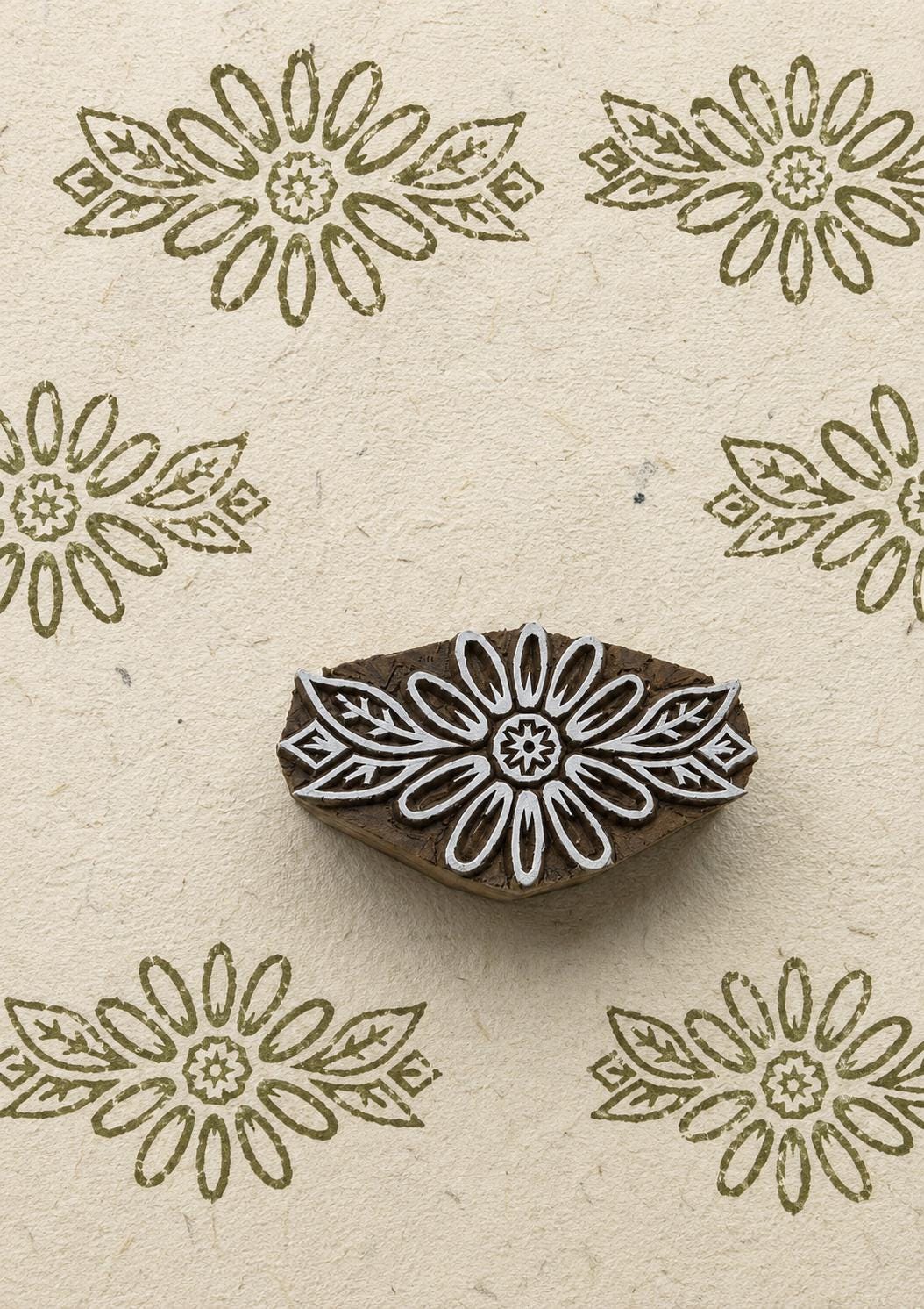 Hand-carved daisy flower wooden printing block surrounded by repeated olive green floral impressions on textured natural handmade paper.