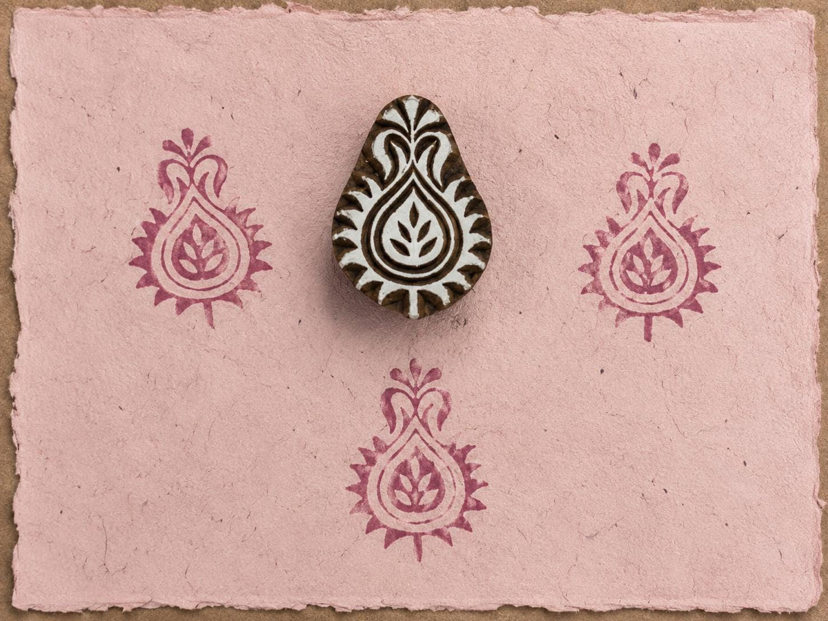 Hand-carved Indian paisley wooden printing block displayed on pink handmade paper with repeating berry-colored paisley impressions.