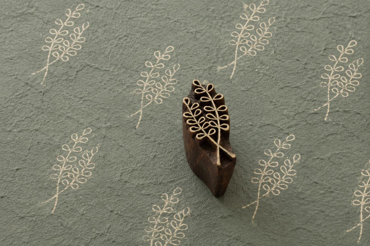 Hand carved wooden printing block featuring a delicate willow fern sprig botanical design stamped repeatedly on textured handmade sage paper.