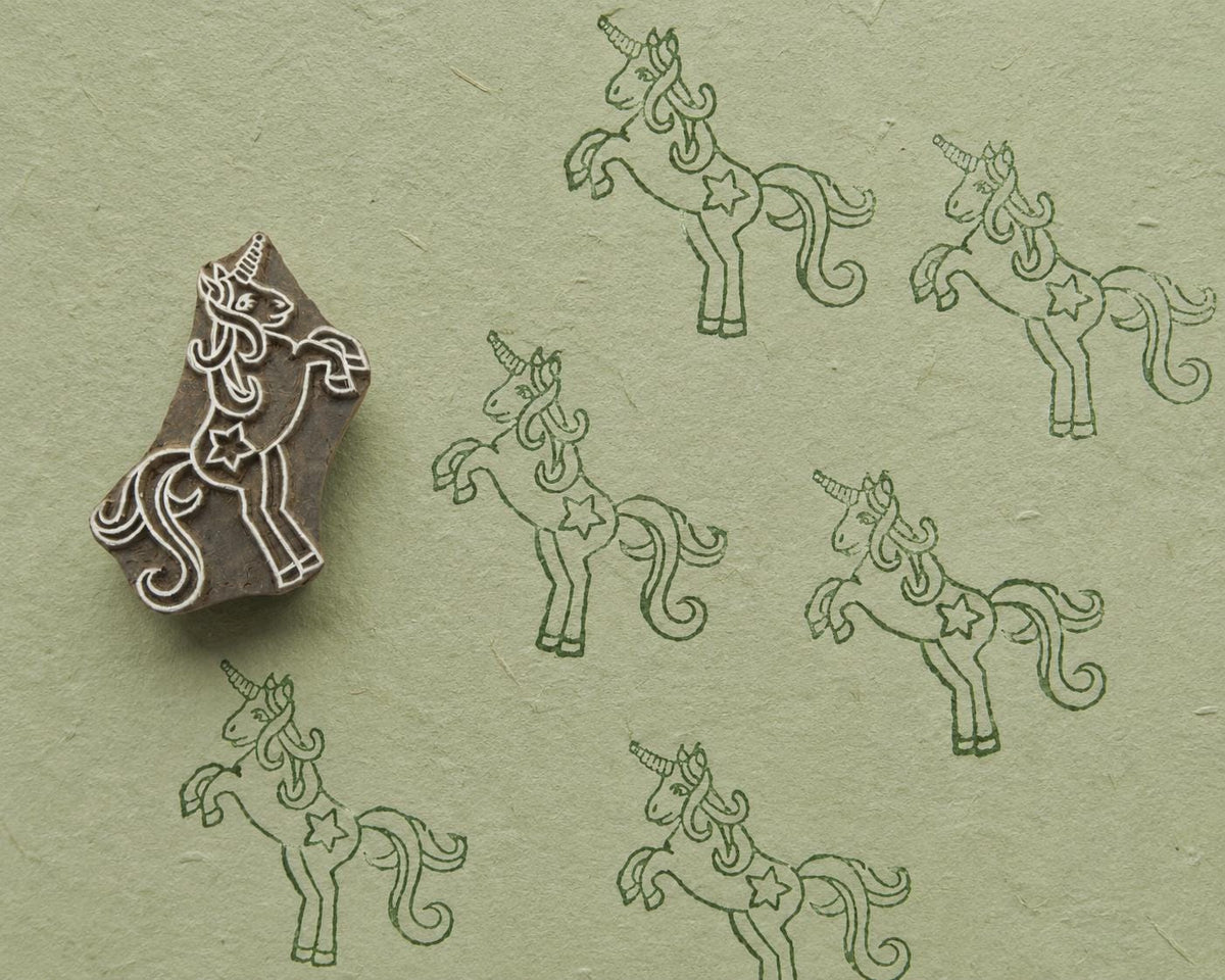Hand carved wooden printing block featuring a prancing unicorn design with repeated dark green impressions on textured sage handmade paper.