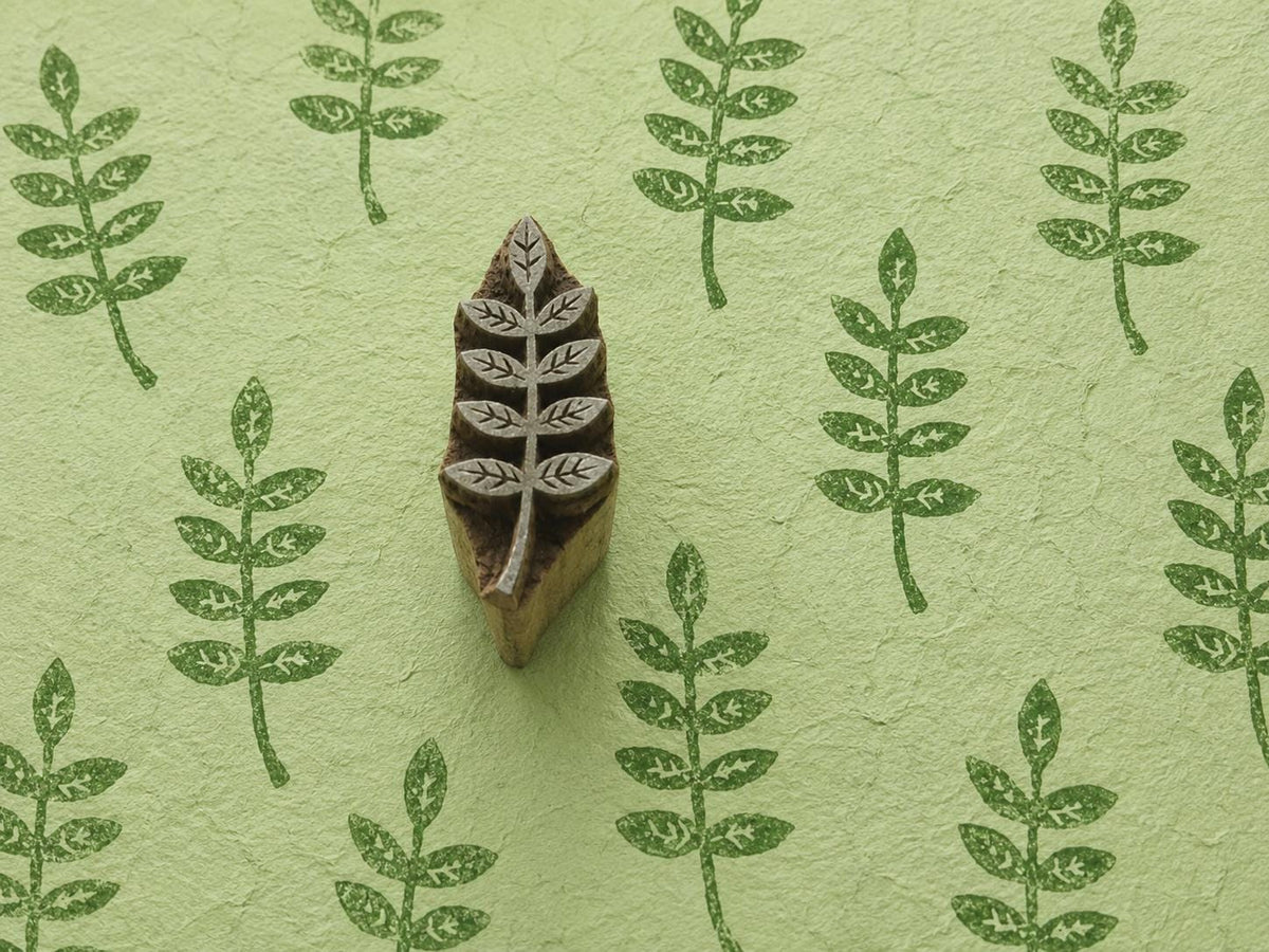 Hand carved wooden leaf printing block displayed on handmade green paper with repeated botanical leaf impressions.