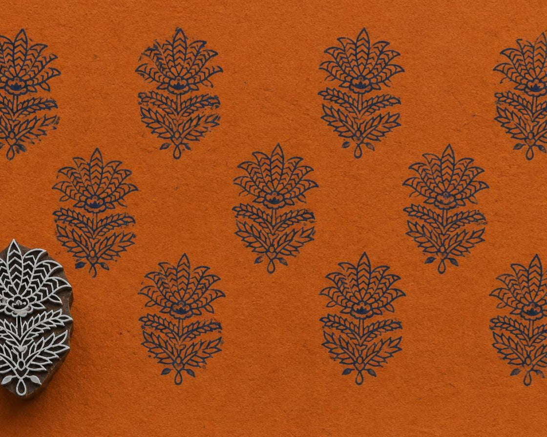Hand-carved Indian floral wooden printing block displayed beside repeated dark blue botanical impressions on textured orange handmade paper.