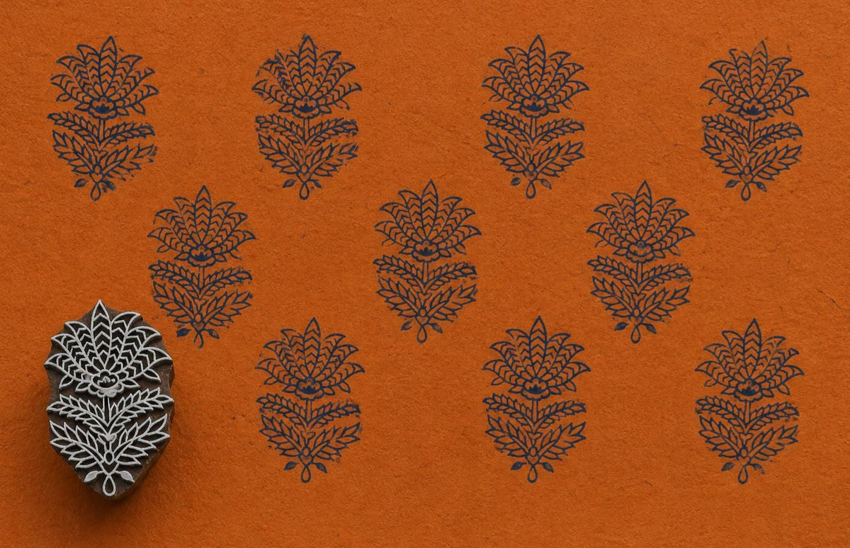 Hand-carved Indian floral wooden printing block displayed beside repeated dark blue botanical impressions on textured orange handmade paper.