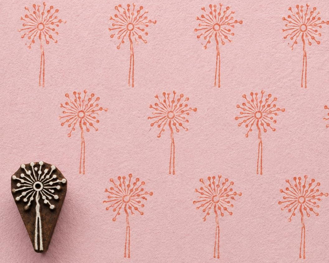 Hand-carved dandelion wooden printing block with repeated coral dandelion impressions on textured pale pink handmade paper.