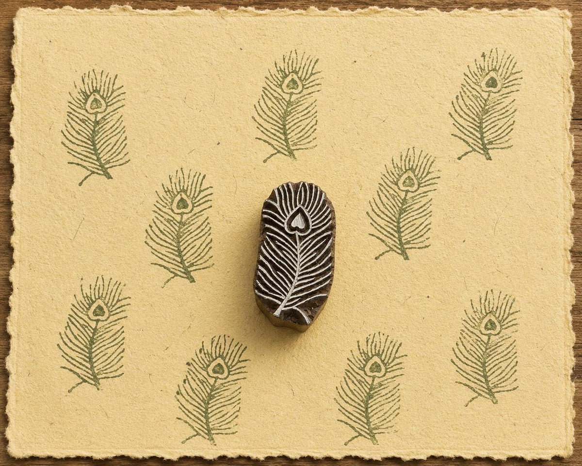 Hand-carved peacock feather wooden printing block surrounded by repeated olive green feather impressions on warm yellow handmade paper.