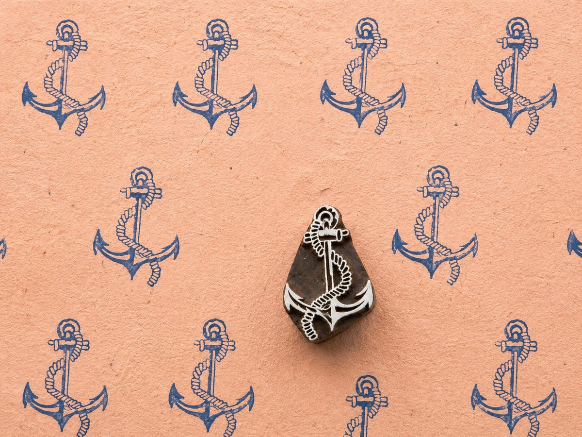 Hand-carved wooden anchor printing block with rope detail surrounded by repeated blue anchor prints on textured peach handmade paper.