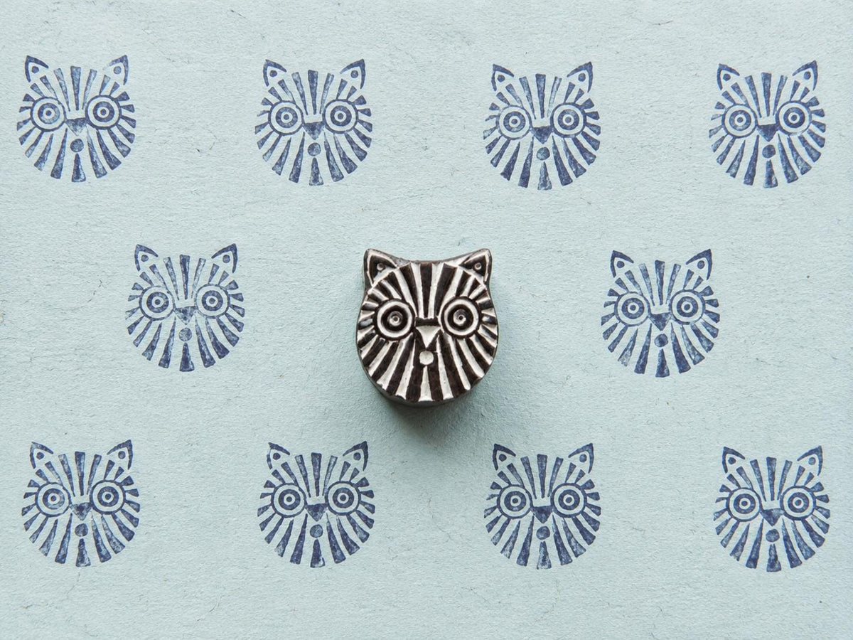Hand-carved tribal cat wooden printing block displayed on pale blue handmade paper with repeating blue geometric cat face impressions.