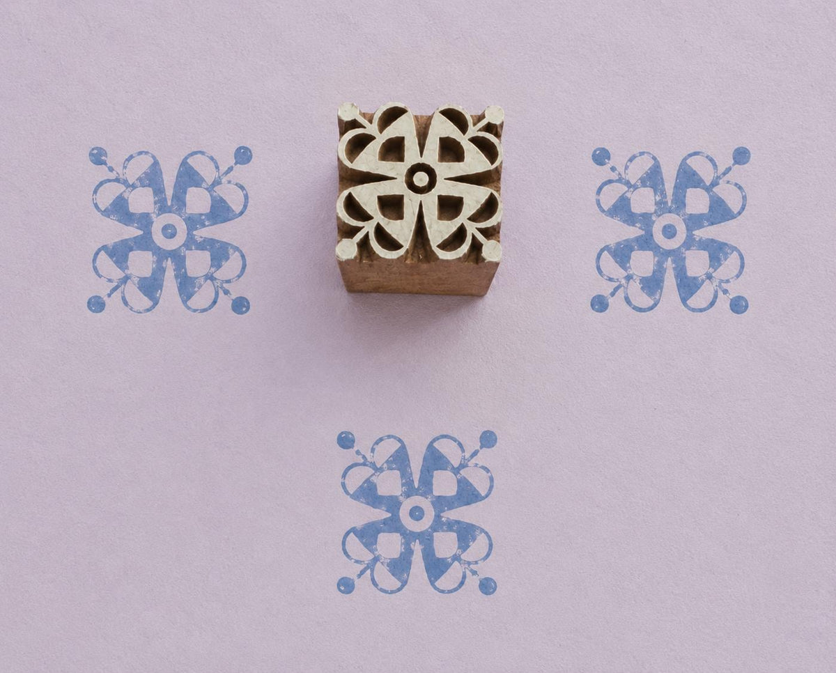 Hand carved wooden printing block featuring a symmetrical Ribbon Bloom motif with looping petals and rounded details, shown with three blue impressions on lavender handmade paper.