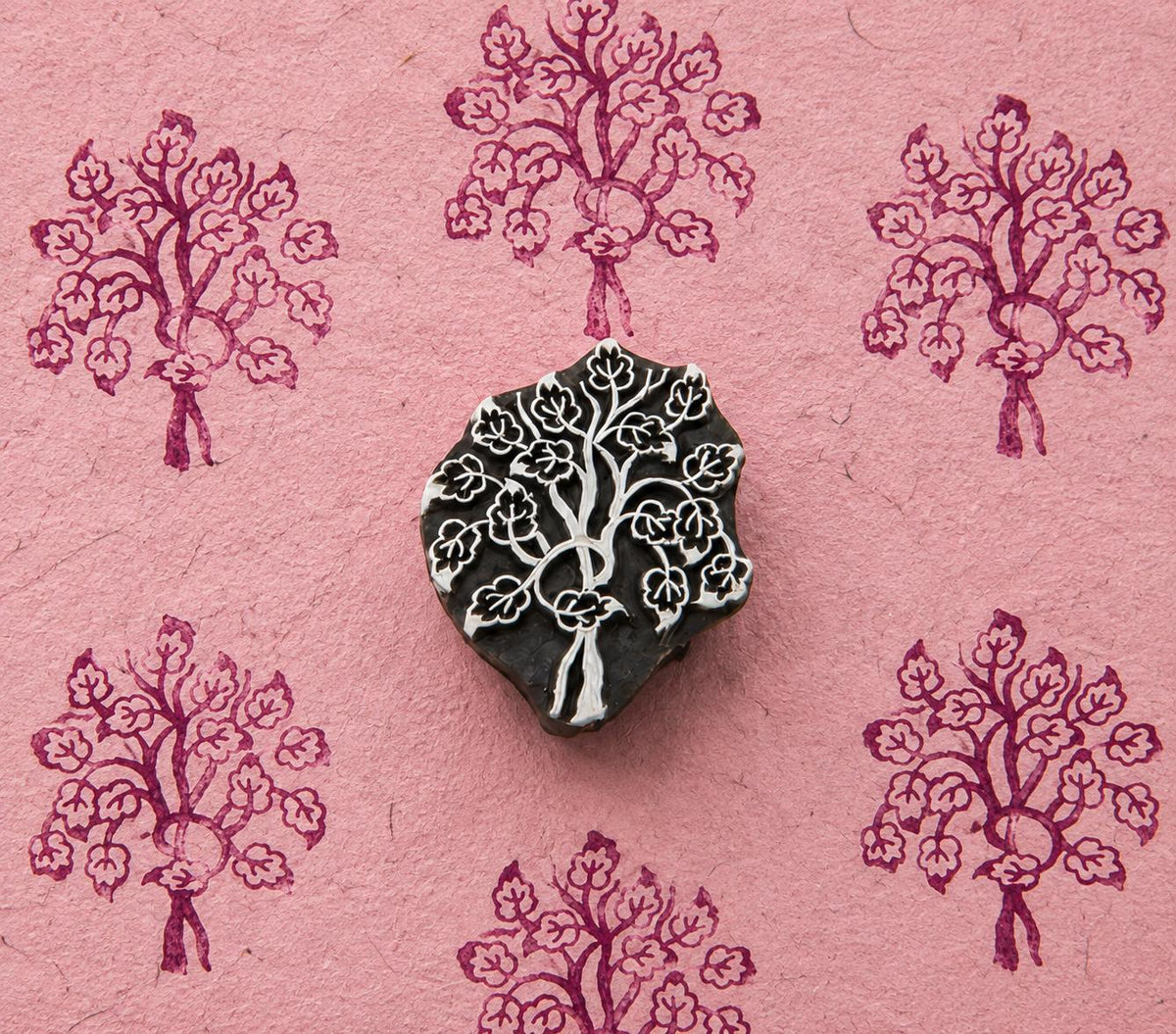 Hand carved wooden printing block featuring a flowering tree motif, surrounded by repeated purple blossom tree impressions on textured pink handmade paper.
