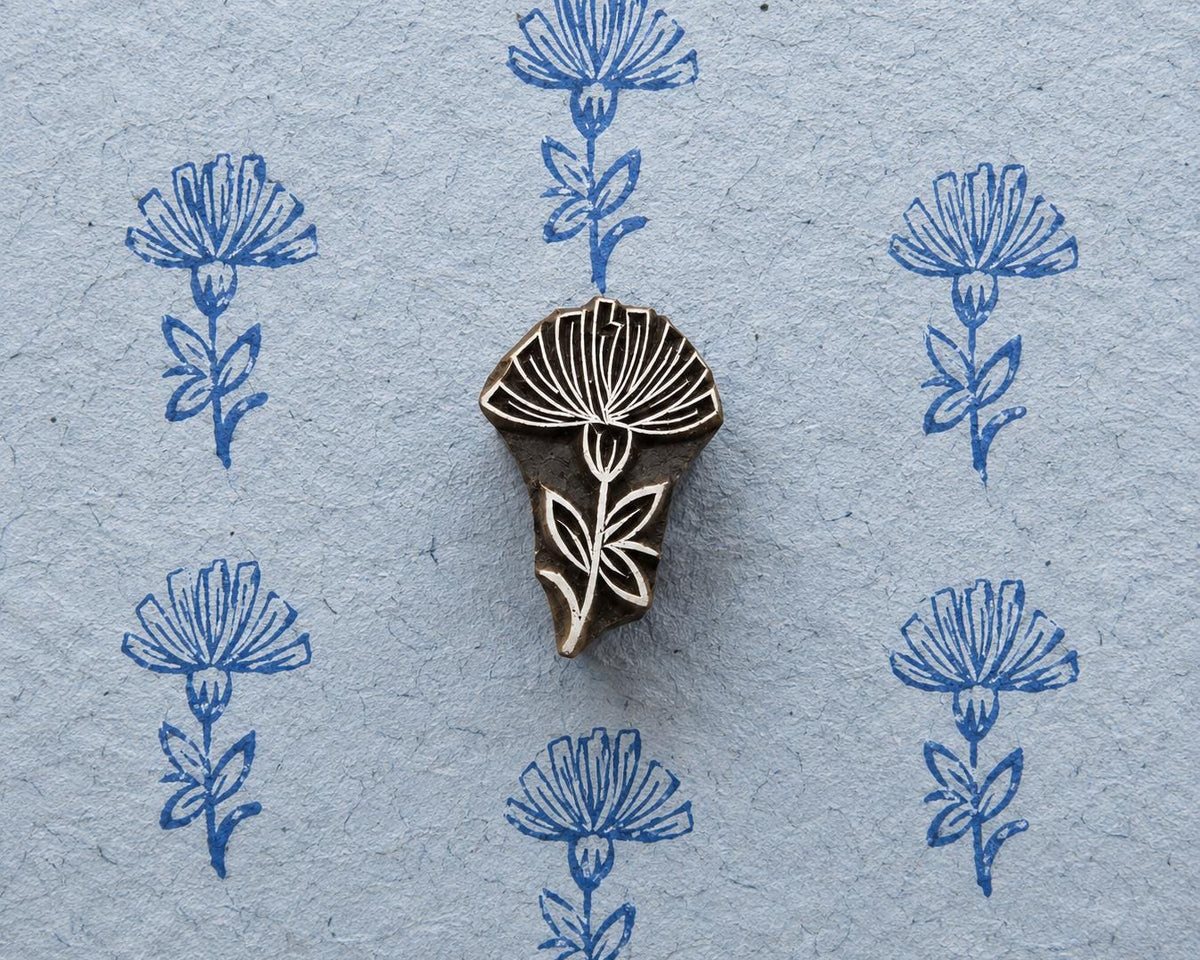 Hand-carved wooden printing block featuring a tall thistle-like flower with a finely detailed bloom, stem and leaves, shown with repeated blue impressions on textured handmade paper.