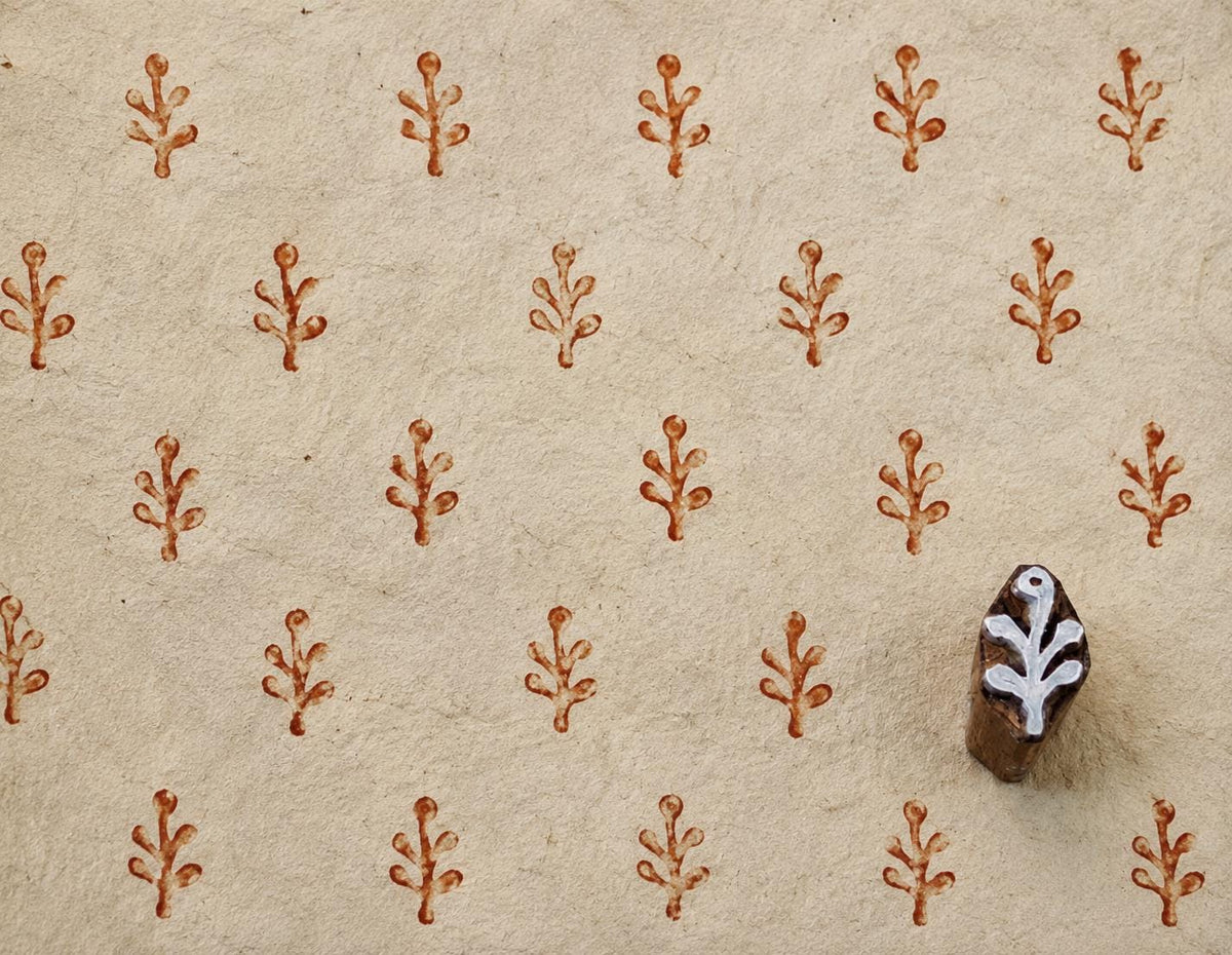 Hand-carved 2 cm wooden printing block with a small branch motif, shown beside repeated reddish-brown impressions on handmade cream paper.