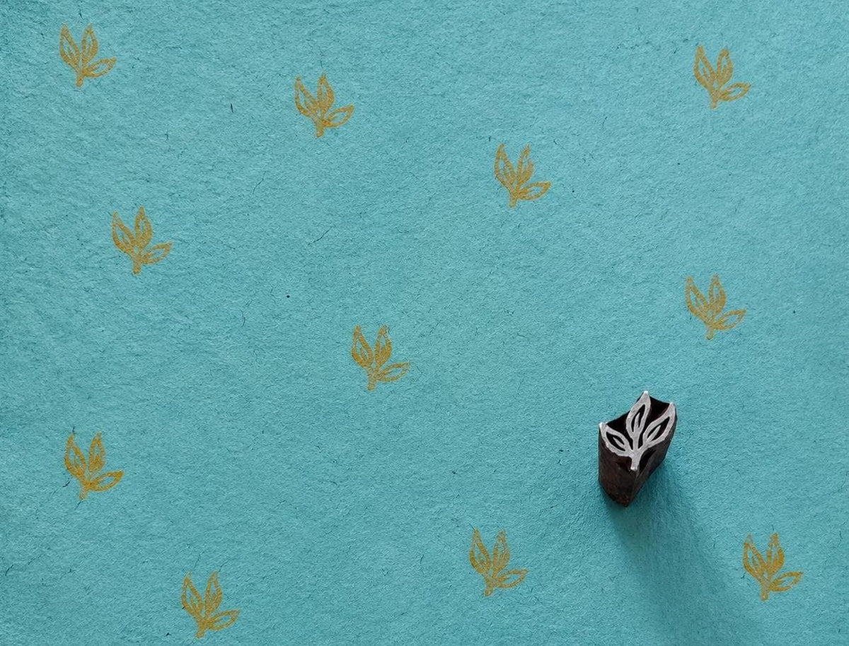 Hand-carved wooden three-leaf sprig printing block beside repeated ochre leaf impressions on textured turquoise handmade paper.