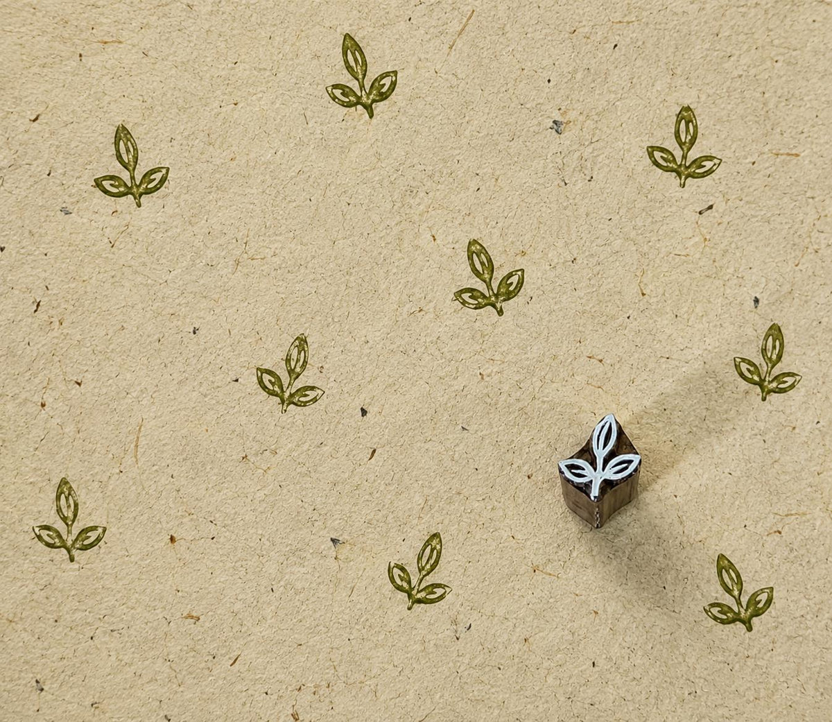 Tiny hand carved wooden printing block with a three-leaf sprout motif, shown with repeated olive green impressions on textured handmade paper