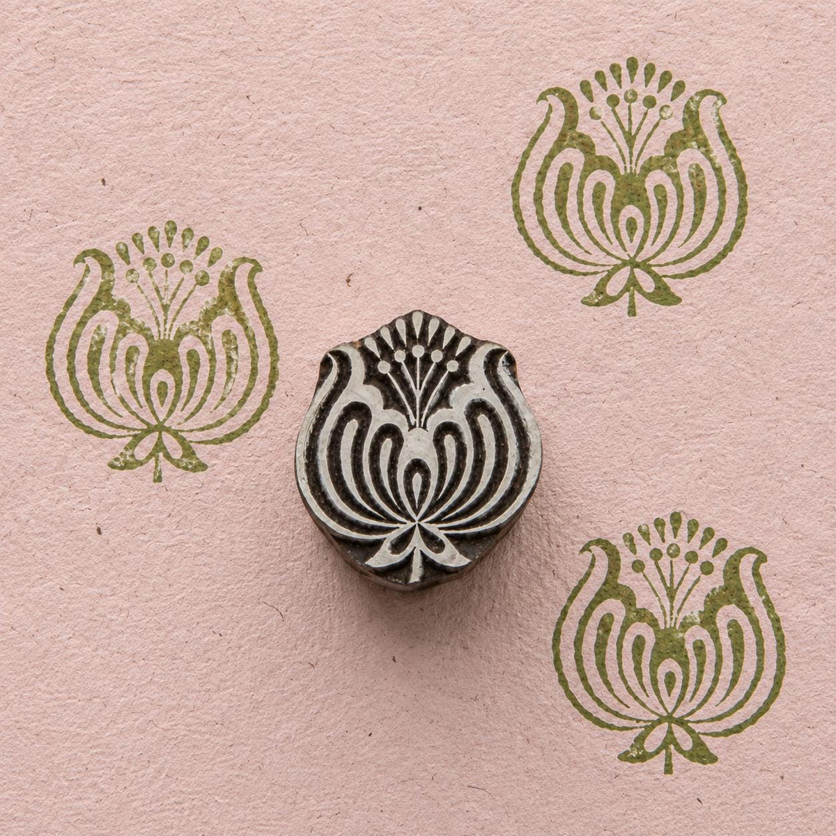 Hand carved wooden ornamental lotus stamp shown with repeated olive green floral impressions on textured pink handmade paper.