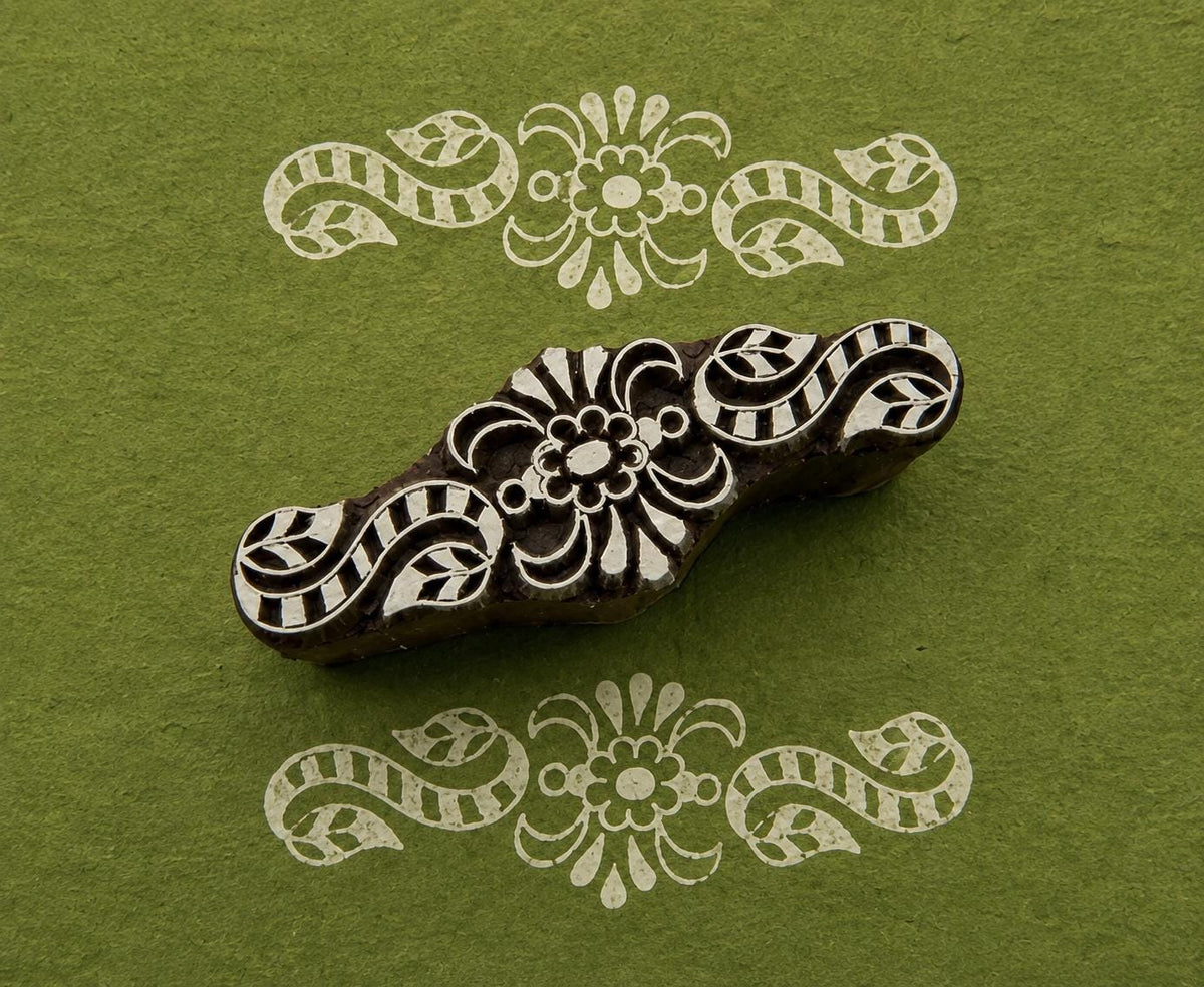 Hand-carved wooden printing block with symmetrical floral scrollwork and curling botanical motifs, shown with white printed impressions on green handmade paper.